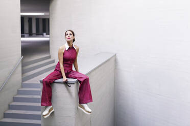 Contemplative woman sitting on wall and looking up near staircase - JSMF02950