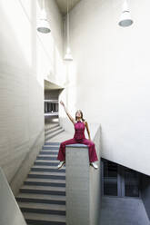 Woman sitting with hand raised on wall near staircase - JSMF02957