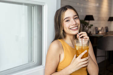 Smiling woman with iced tea leaning on door at home - DSIF00745