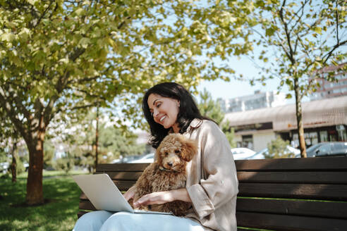 Smiling freelancer using laptop with poodle dog sitting on lap at park - MDOF01685