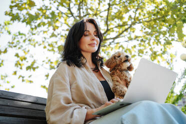 Smiling freelancer holding poodle dog and working on laptop at park - MDOF01690