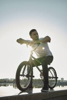 Nachdenklicher Mann sitzt auf BMX-Rad in der Nähe von See unter Himmel - MRPF00027