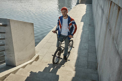 Lächelnder Mann mit BMX-Rad in der Nähe eines Sees auf einer Böschung stehend - MRPF00045