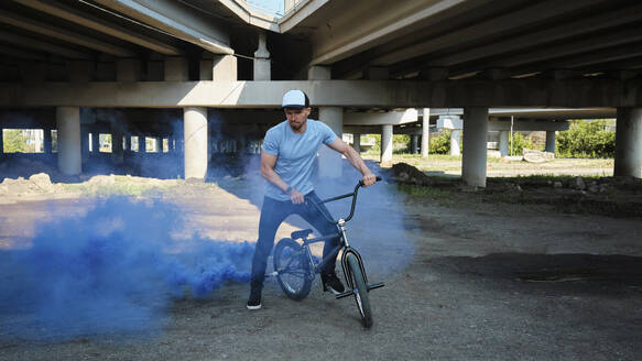 Mann mit BMX-Rad stößt in der Nähe einer Brücke blauen Rauch aus - MRPF00050