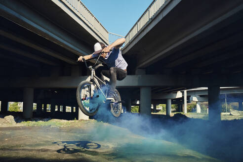 Mann springt mit BMX-Rad und stößt in der Nähe einer Brücke blauen Rauch aus - MRPF00053