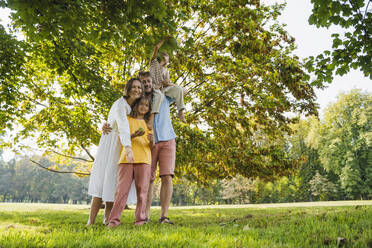 Glückliche Familie unter einem Baum im Park stehend - NDEF01422