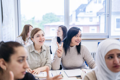 Lächelnder Schüler, der neben einem Freund im Klassenzimmer sitzt und das Friedenszeichen macht - NJAF00627