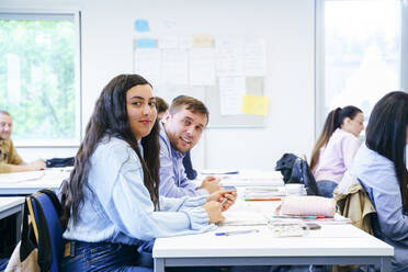 Lächelnde verschiedene Freunde sitzen am Schreibtisch mit Büchern im Klassenzimmer - NJAF00631