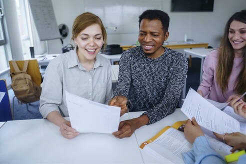 Lächelnde multiethnische Studenten mit Notizen, die gemeinsam am Schreibtisch im Klassenzimmer lernen - NJAF00667