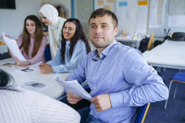Nachdenklicher Mann mit Papier in der Hand sitzt mit multiethnischen Schülern im Klassenzimmer - NJAF00673