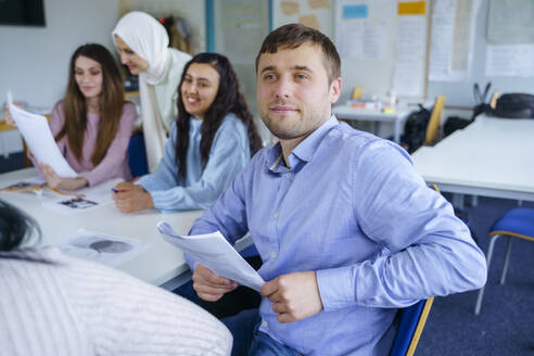 Nachdenklicher Mann mit Papier in der Hand sitzt mit multiethnischen Schülern im Klassenzimmer - NJAF00673