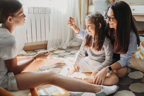 Smiling mother and daughters playing rock paper scissors on carpet in living room - ANAF02408