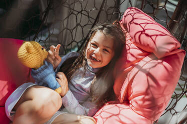 Lächelndes Mädchen mit Plüschtier auf Veranda-Schaukel liegend - ANAF02420
