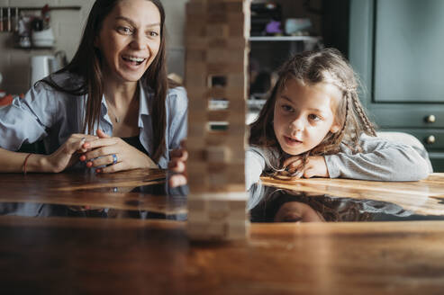 Smiling mother watching daughter playing block removal game at table - ANAF02434