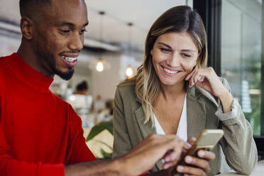 Lächelnde Geschäftsfrau, die mit einem Kollegen in einem Café sitzt und ein Smartphone benutzt - JOSEF22095