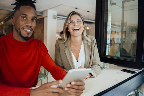 Lachende Geschäftsfrau, die mit einem Kollegen in einem Café sitzt und einen Tablet-PC hält - JOSEF22132
