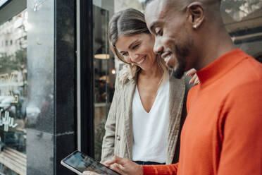 Lächelnde Geschäftsfrau mit jungem Kollegen, der vor einem Café einen Tablet-PC benutzt - JOSEF22134