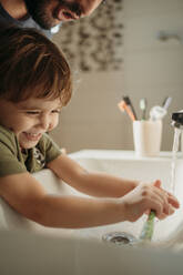 Man with happy boy washing tooth brush in sink at home - ANAF02457