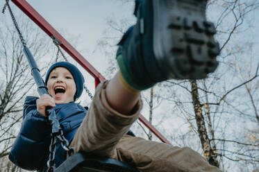 Cheerful boy playing on swing at park under sky - ANAF02460