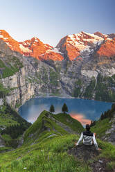 View of a hiker resting in front of the Oeschinensee lake surrounded by snowy peak at sunset, Oeschinensee, Kandersteg, Bern Canton, Switzerland, Europe - RHPLF29066