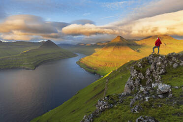 Summer view of a hiker standing on top of a mountain overlooking the fjord at sunrise, Funningur fjord, Eysturoy island, Faroe islands, Denmark, Europe - RHPLF29070