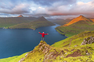 Happy hiker stands on top of the cliff overlooking the fjord of Funningur, Eysturoy island, Faroe islands, Denmark, Europe - RHPLF29074