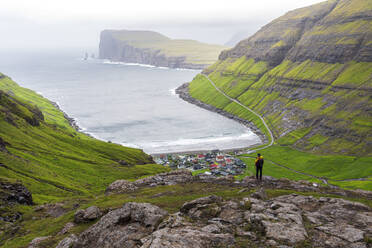 One man on top of a rock admiring the village of Tjornuvik in the misty weather, Sunda municipality, Streymoy island, Faroe islands, Denmark, Europe - RHPLF29076