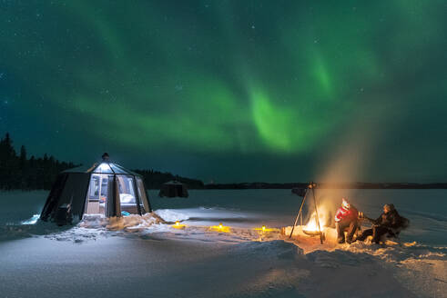 Night view of two people enjoyng dinner outside around a campfire close to the illuminated glass igloo with Northern Lights (Aurora Borealis) dancing in the sky, Jokkmokk, Norrbotten, Swedish Lapland, Sweden, Scandinavia, Europe - RHPLF29084