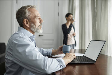 Man using laptop and drinking tea while spending time at home with wife - KPEF00360