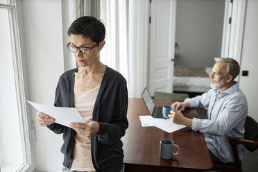 Pensive woman looking at business paper standing next to husband sitting at table working on laptop - KPEF00362