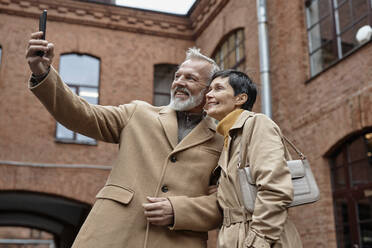 Happy elderly couple taking selfie on smartphone during city walk - KPEF00384