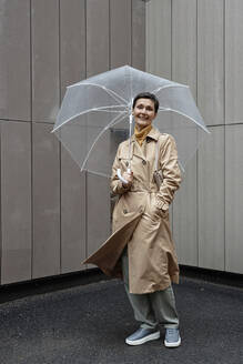 Glückliche Frau in beigem Regenmantel, die einen Regenschirm hält, während sie draußen spazieren geht und in die Kamera schaut - KPEF00393