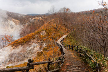 Wanderweg durch einen herbstlichen Wald mit dem dampfenden Vulkantal des Noboribetsu auf der linken Seite, Hokkaido, Japan, Asien - RHPLF29483