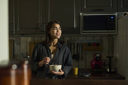 Smiling woman holding bowl of porridge and standing in kitchen at home - DSHF01132