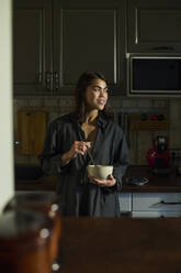 Lächelnde Frau, die eine Schüssel mit Frühstück hält und zu Hause in der Küche steht - DSHF01133