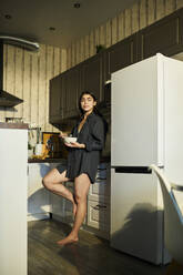 Nachdenkliche Frau, die sich auf den Küchentisch stützt und eine Schüssel mit Frühstück in der Hand hält - DSHF01138
