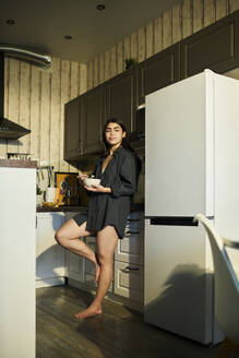 Thoughtful woman leaning on kitchen counter and holding bowl of breakfast at home - DSHF01138