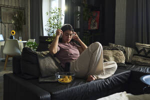 Frau trägt drahtlose Kopfhörer und sitzt mit einer Schüssel mit Snacks auf dem Sofa zu Hause - DSHF01144