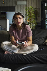 Woman wearing wireless headphones and holding game controller on sofa at home - DSHF01147