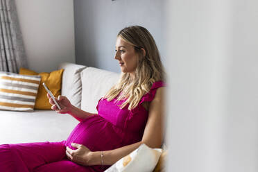 Lächelnde schwangere Frau, die auf dem Sofa sitzt und ihr Smartphone zu Hause benutzt - WPEF07841