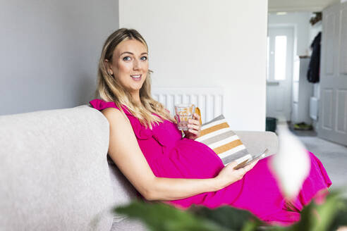 Glückliche schwangere Frau sitzt mit Smartphone und einem Glas Wasser auf dem Sofa - WPEF07843