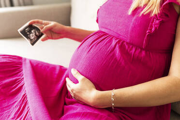 Pregnant woman holding ultrasound photograph with hand on stomach at home - WPEF07846