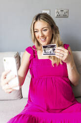 Lächelnde schwangere Frau zeigt Ultraschallfoto auf Videoanruf durch Smartphone zu Hause - WPEF07847