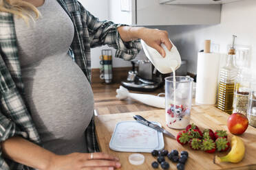 Pregnant woman pouring milk in measuring cup and preparing milkshake - WPEF07854