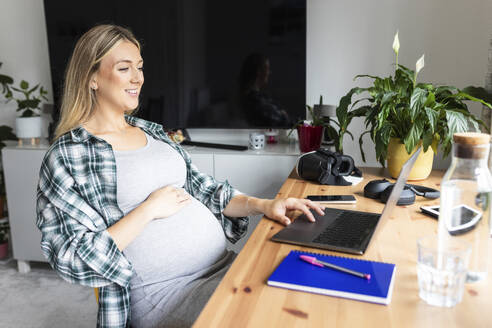 Lächelnde schwangere Freiberuflerin bei der Arbeit am Laptop im Home Office - WPEF07864