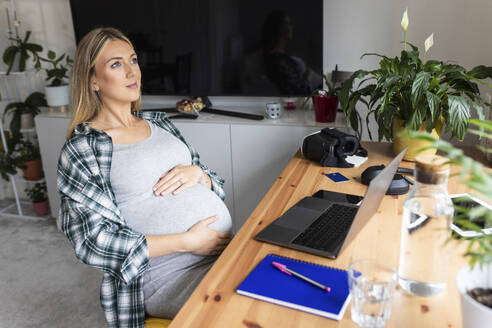 Contemplative pregnant freelancer sitting with laptop at desk in home office - WPEF07866
