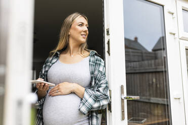 Lächelnde schwangere Frau, die ein Smartphone hält und in der Nähe der Balkontür steht - WPEF07869