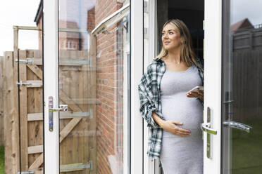 Lächelnde schwangere Frau, die mit der Hand auf dem Bauch steht und das Smartphone an der Balkontür hält - WPEF07870