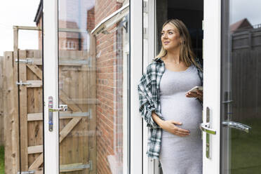 Smiling pregnant woman standing with hand on stomach and holding smart phone at balcony door - WPEF07870