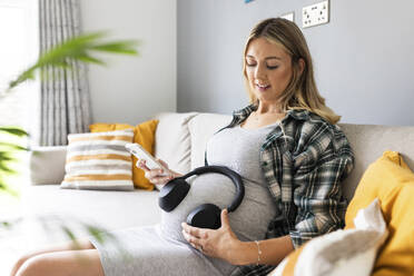 Smiling pregnant woman sitting with smart phone and wireless headphones on stomach at home - WPEF07871